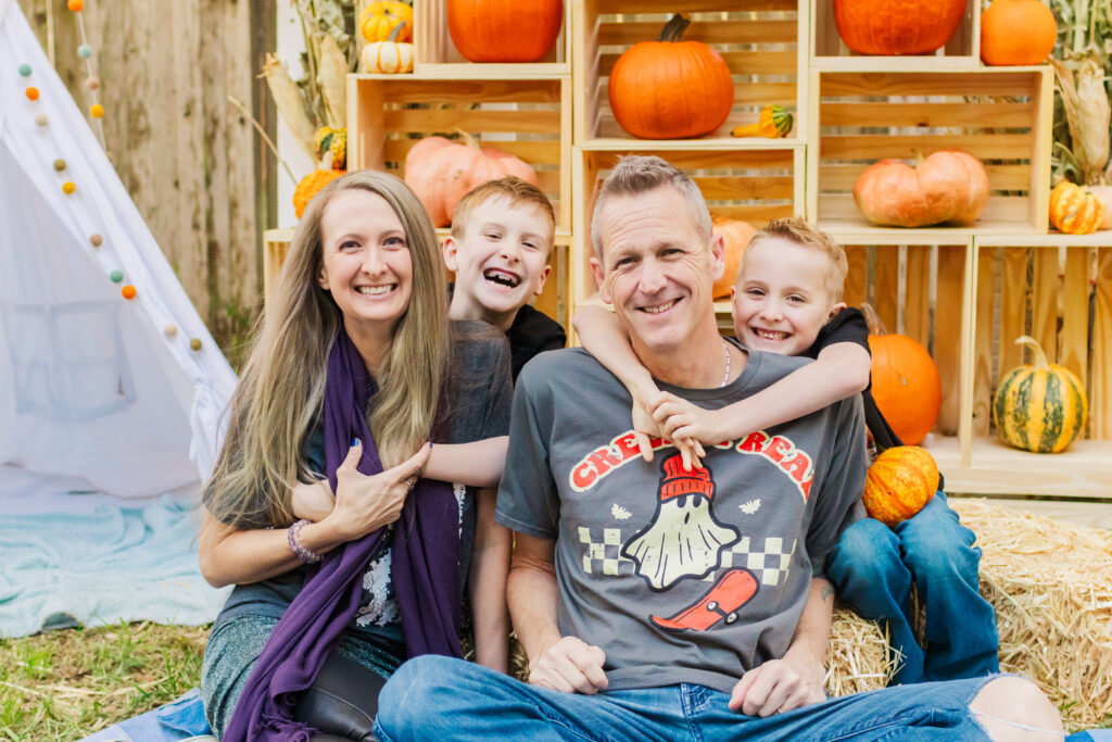 A family of four sits together at a local San Antonio pumpkin patch, posted in front of a Fall themed background, with two boys hugging their parents from behind, surrounded by wooden crates full of pumpkins and gourds.