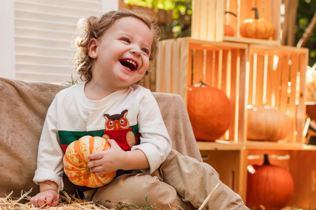 A laughing toddler dressed in a cute Fall outfit holds a small striped gourd while sitting on hay bales in front of a Fall-themed background decorated with pumpkins.